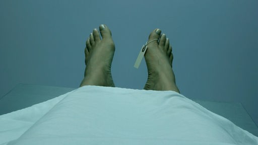 Nackte Füße unter einem Laken. Am rechten großen Zeh hängt ein Zettel