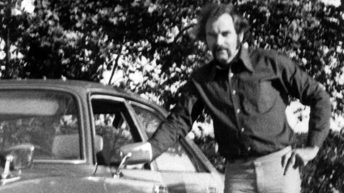 Schwarzweiß-Bild aus den 70er Jahren. Ein Mann lehnt lässig an der Seite eines Autos.