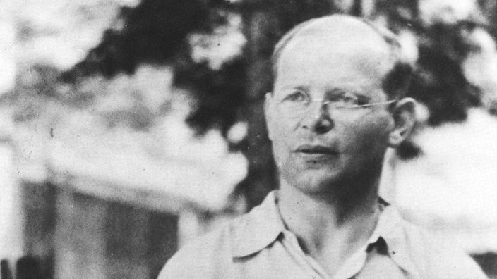 Schwarz-Weiß-Aufnahme von Bonhoeffer auf dem Hof des Wehrmachtgefängnisses. In seiner rechten Hand hält er ein Buch.