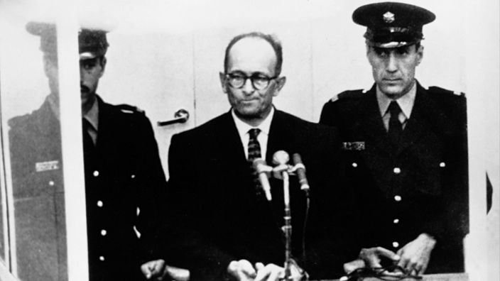 Adolf Eichmann steht hinter Glasscheiben an einem Mikrofon, links und rechts neben ihm zwei uniformierte Männer