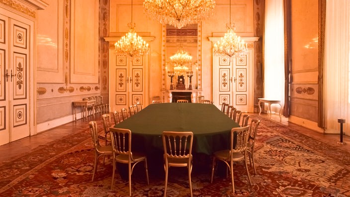 Ein runder, langer Tisch mit vielen Stühlen in einem prachtvoll geschmückten Saal mit Kronleuchtern
