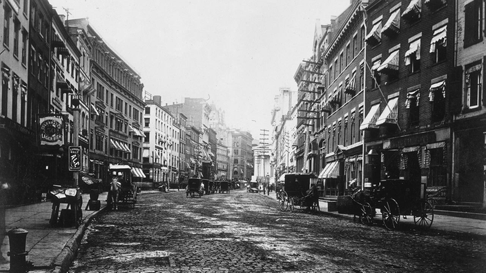 Die Schwarzweiß-Fotografie aus dem Jahr 1882 zeigt eine Straße mit Kopfsteinpflaster. Links und rechts stehen viergeschossige Häuser, davor Kutschen und Pferdefuhrwerke.