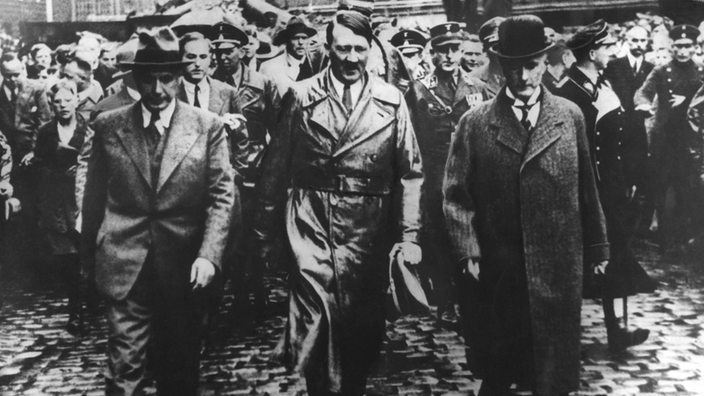 Adolf Hitler (Mitte) und Gustav Krupp von Bohlen und Halbach (rechts) gehen über eine Straße des Krupp-Konzerns; hinter ihnen sind Polizisten und Zuschauer.