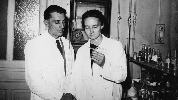 Frederic Joliot-Curie und seine Frau Irene Joliot Curie sind in einem Labor zu sehen. Sie tragen weiße Kittel und betrachten einen Gegenstand, den Irene in der Hand hält.