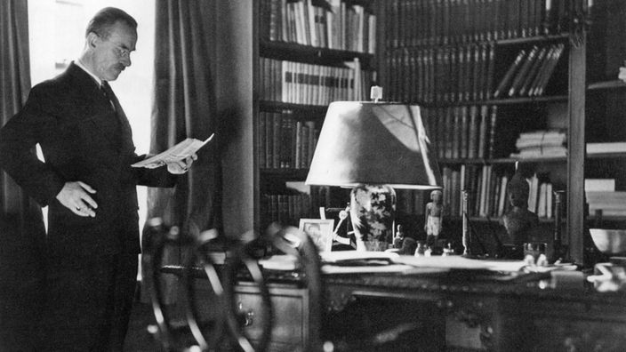 Schwarzweiß-Foto: Thomas Mann vor seinem Schreibtisch stehend, in der Hand ein Blatt Papier, im Hintergrund Bücherwände.