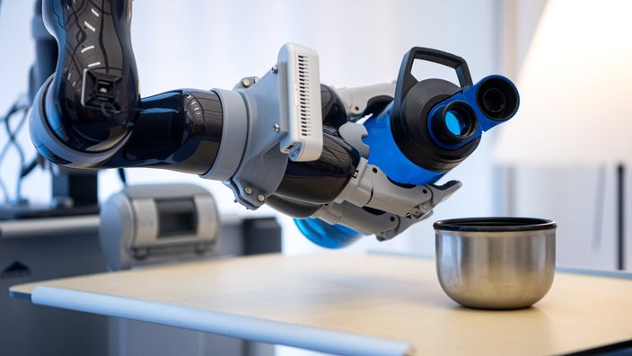 Ein Roboterarm gießt bei einem Versuchsaufbau am Deutschen Forschungszentrum für Künstliche Intelligenz Wasser in einen Becher. 