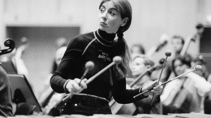 Das Bild zeigt eine junge Frau mit langen Haaren und Zopf. Sie sitzt hinter einem Schlagzeug, hinter ihr sind viele Cellisten zu sehen. Sie schaut zur Seite.