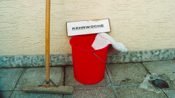 Ein Schrubber lehnt gegen eine Hauswand, daneben steht ein roter Eimer, auf dem ein Schild mit der Aufschrift "Kehrwoche" liegt.
