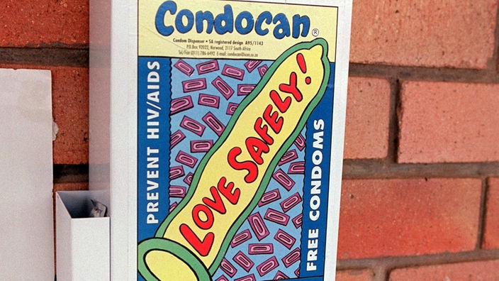 An einer roten Backstein-Hauswand hängt ein weißer Kondomautomat mit bunter Aufschrift und Aids-Schleife, in dessen Fach versiegelte Kondome liegen. Eine der Aufschriften bedeutet übersetzt 'Greif zu - kostenlose Kondome'.