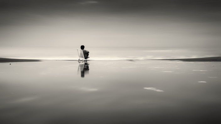 Eine Frau sitzt mit angezogenen Beinen auf einem Stuhl, der im Meer steht und schaut auf das Wasser. 
