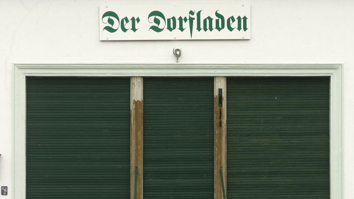 Blick auf einen geschlossenen Laden über dessen Eingangstür ein Schriftzug 'Der Dorfladen' prangt.