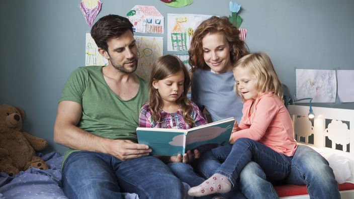 Ein Ehepaar sitzt im Kinderzimmer auf dem Bett und blättert mit zwei Töchtern in einem Buch.