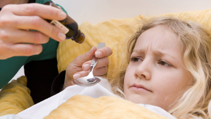 Ein im Bett liegendes Mädchen schaut skeptisch auf einen Löffel vor sich, auf den eine Person Medizin tropfen lässt.