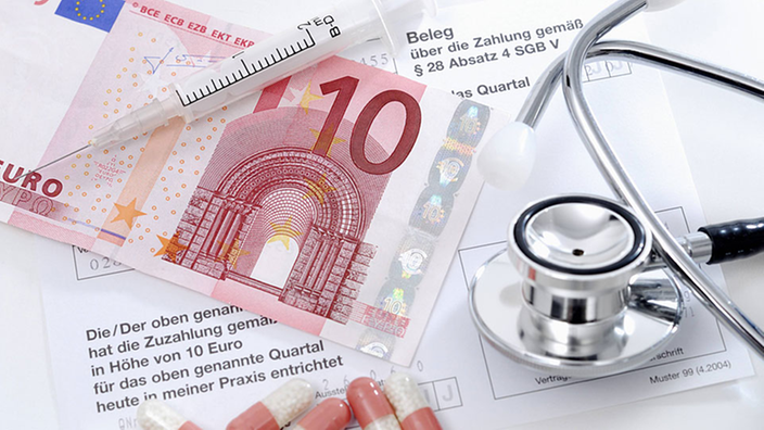 Symbolfoto mit einem Zehn-Euro-Schein, einem Stethoskop, einer Quittung und ein paar Medikamentenkapseln.