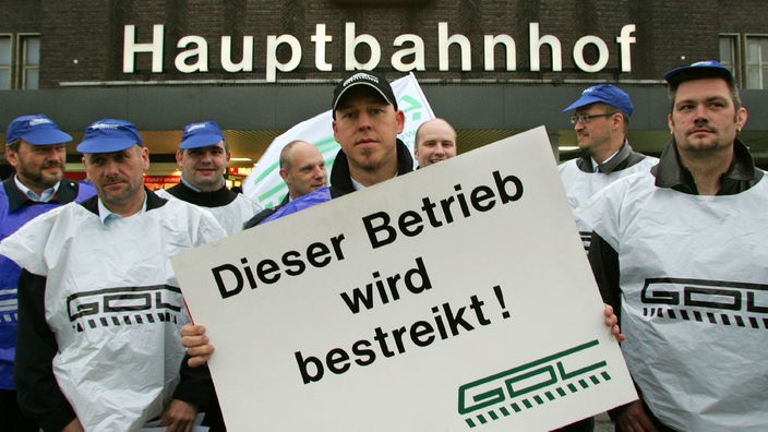 Streikende Lokführer halten vor einem Hauptbahnhof ein Schild mit der Aufschrift "Dieser Betrieb wird bestreikt. GDL" hoch.