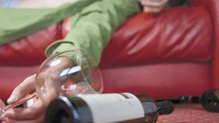 Ein Mann liegt auf einem roten Sofa und hat einen Arm über das Gesicht gelegt. In der anderen Hand hält er ein leeres Weinglas. Am Boden liegt eine Weinflasche.