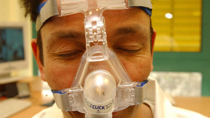 Großaufnahme vom Gesicht eines schlafenden Mannes. Über der Nase liegt eine durchsichtige Atemmaske. Diese ist mit Halterungsgurten über Stirn und Wangen befestigt.