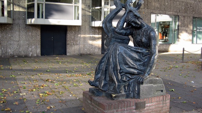 Die Skulptur der Dichterin Sappho steht auf dem Kölner Offenbachplatz, Herbstlaub liegt auf dem Boden.