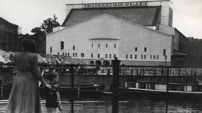 Schwarz-weiß-Fotografie des Friedrichstadt-Palastes aus dem Jahr 1949. Totale. Vor dem Gebäude stehen eine Frau und ein Junge mit dem Rücken zur Kamera und schauen auf das Revue-Theater.