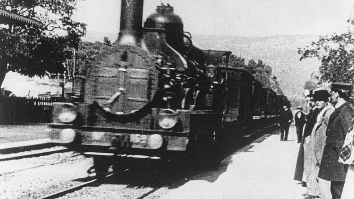 Schwarzweiß-Aufnahme einer fahrenden Dampflokomotive. Am Bahnsteig stehen Menschen.
