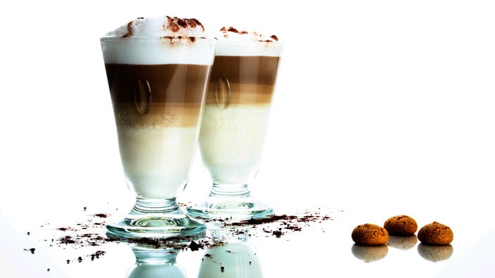 Zwei Gläser mit einer kunstvoll gestalteten Milchkaffee-Mischung: Unten im Glas ist Milch, es folgen zwei Schichten Kaffee und Milchschaum. Um die Gläser sind Schokostreusel drapiert, daneben liegen kleine, runde Mandelkekse.