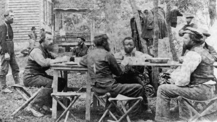 Eine Schwarzweiß-Fotografie aus dem Jahr 1862. Männer sitzen zur Besprechung an einem Tisch im Freien