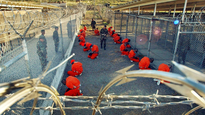 18 Gefangene in orangefarbenen Overalls knien auf einem offenen Platz, hoch mit Stacheldraht umzäunt, daneben fünf bewaffnete US-Militärpolizisten.
