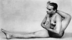 Schwarzweiß-Aufnahme von B.K.S. Iyengar, der auf dem Boden sitzt, ein Bein ausgestreckt und das andere Bein nach hinten gedehnt hat, so dass sich ein Fuß hinter seinem Kopf befindet