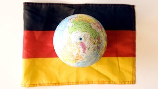 Eine Deutschlandfahne liegt auf weißem Untergrund; auf ihr ruht ein Globus.