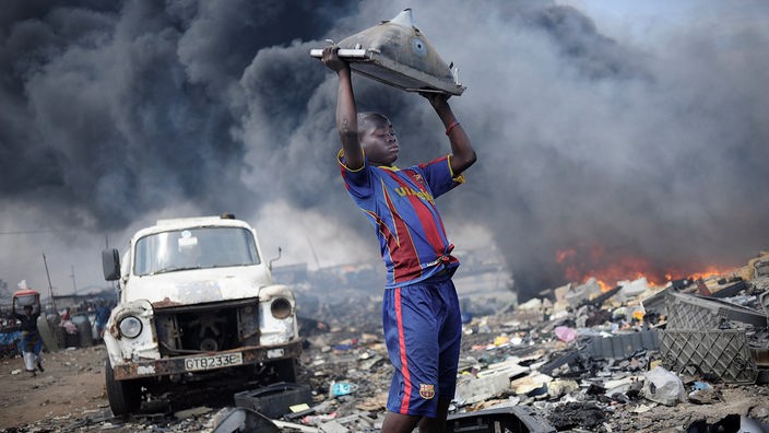 Ein Junge hebt auf der Giftmüllhalde Agbogbloshie in Ghana einen alten Monitor nach oben, hinter ihm toxischer Rauch.