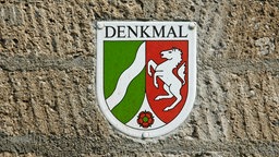 In einer alten Mauer ist eine Plakette eingelassen, auf der der Schriftzug Denkmal sowie das Wappen von NRW zu sehen sind. | Bildquelle: WDR / Interfoto In einer alten Mauer ist eine Plakette eingelassen, auf der der Schriftzug Denkmal sowie das Wappen von NRW zu sehen sind.