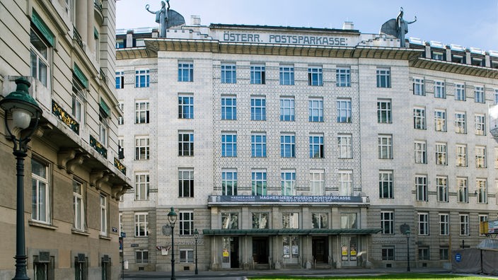 Ein Steinbau mit fünf Fensterreihen an dem oben der Schriftzug Österreichische Postsparkasse prangt. Die Fassade des Gebäudes schmücken nur reihenförmig angeordnete Knöpfe aus Stein 
