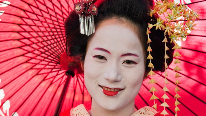 Porträt einer Geisha in rotem Kimono. Sie trägt einen roten Schirm. 