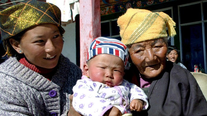 Drei Tibeter in Tracht: Links eine junge Frau, in der Mitte ein Baby, rechts eine sehr alte Frau.