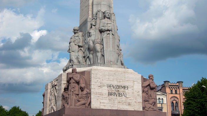 Das Foto zeigt das Freiheitsdenkmal in Riga. Am Fuß des Monolithen aus Stein sind verschiedene Figuren abgebildet