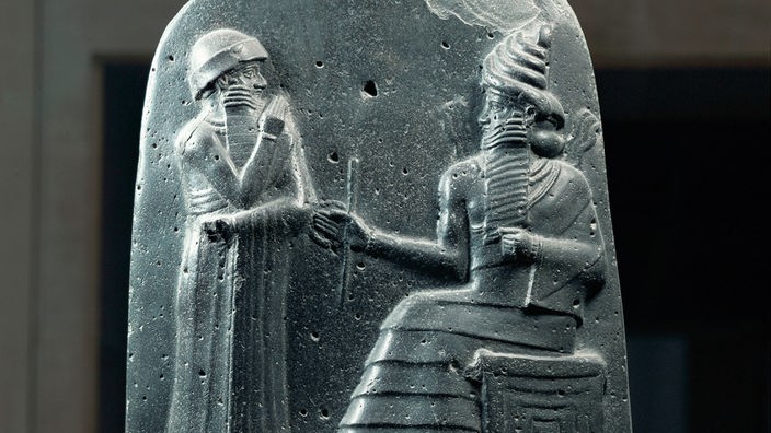 Ein Steinrelief aus vorchristlicher Zeit zeigt den König Hammurabi in sitzender Position auf einem Thron.