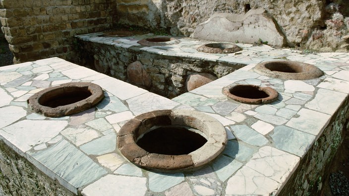 Eine aus grobem Stein gemauerte Theke in eckiger U-Form. In die Theke eingelassen sind runde Muldenöffnungen, die in römischer Zeit als Feuerstelle in Garküchen dienten.