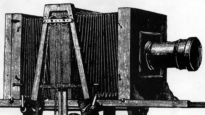 Diese Zeichnung zeigt eine Plattenkamera aus dem Jahr 1866, neben der ein Fotograf steht. Das Erstaunliche ist der Größenvergleich: Die Kamera ist inklusive Stativ fast doppelt so groß wie der Fotograf.