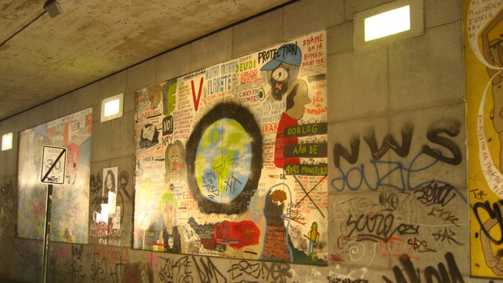 Die grauen Betonwände der Unterführung werden von einigen großflächigen bunten Bildern verschönert, die ihrerseits wiederum teilweise mit Graffiti bedeckt sind.