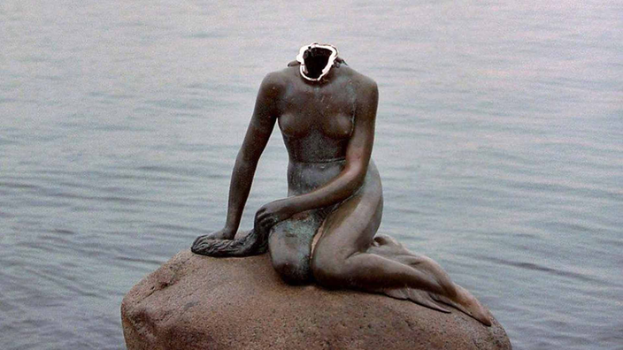 Die Bronzefigur sitzt mit abgeschlagenem Kopf auf ihrem Stein im Wasser.
