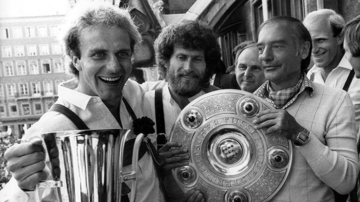 Karl-Heinz Rummenigge, Paul Breitner und Pal Csernai halten bei der Meisterschaftsfeier 1981 einen Pokal und die Schale des Deutschen Meisters in die Kamera.
