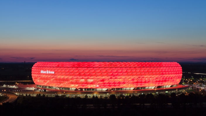 Die ovale Arena in der Abenddämmerung. Die rautenförmigen Luftkissen sind rot beleuchtet, so dass das ganze Stadion von außen betrachtet wie eine rote Kuppel aussieht.
