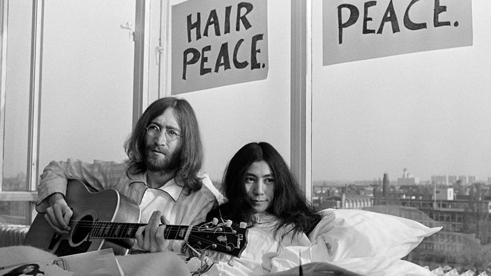 John Lennon und Yoko Ono sitzen in einem Bett, über ihnen zwei Schilder mit den Aufschriften "Hair Peace" und "Bed Peace"