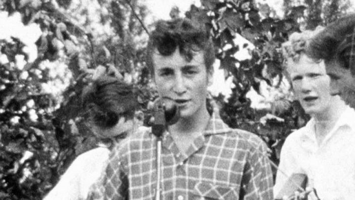 John Lennon als Jugendlicher mit hochtoupierten Haaren spielt Gitarre und singt in ein Mikrophon.