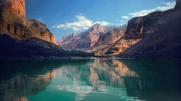 Die rötlichen Felsen des Grand Canyon. Im Vordergrund das türkisblaue Wasser des Colorado Rivers, in dem sich die Felsen spiegeln.