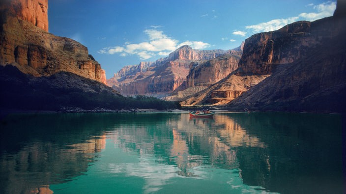 Die rötlichen Felsen des Grand Canyon. Im Vordergrund das türkisblaue Wasser des Colorado Rivers, in dem sich die Felsen spiegeln.