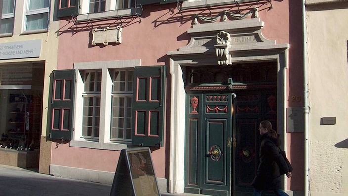 Das Foto zeigt Beethovens Geburtshaus in Bonn. Der mehrstöckige Altbau, an den links und rechts andere Häuser angrenzen, ist rosa gestrichen mit dunkelgrünen Fensterläden und einer dunkelgrünen Tür sowie einem dunklen Schieferdach. Vor dem Haus steht ein Plakatständer. Eine dunkel gekleidete Person geht am Haus vorbei. Die Sonne scheint auf das Haus.