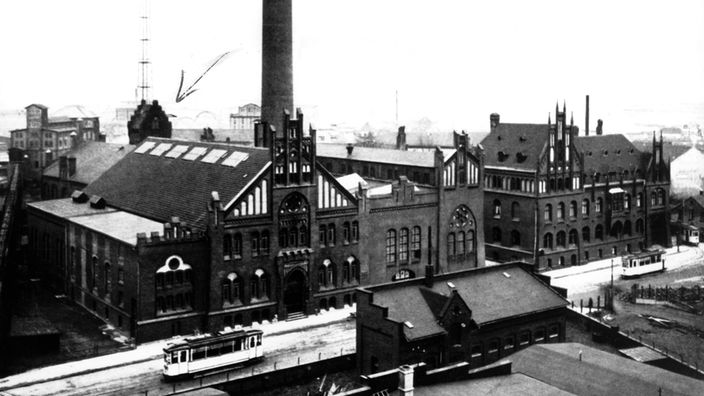 Ein altes Schwarzweiß-Foto zeigt ein Fabrikgelände an einer von Straßenbahnen befahrenen Hauptstraße. In einem der Gebäude befand sich das Funkhaus Münster, im Hintergrund links kann man den Sendemast erkennen.