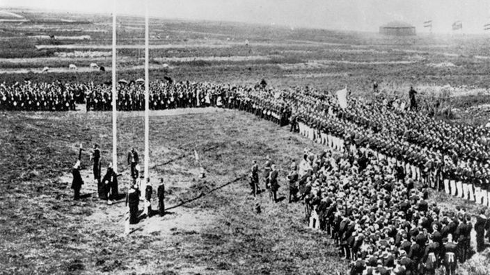 Das Bild zeigt eine Luftaufnahme eines großen Platzes auf dem hunderte Soldaten versammelt sind. In der Mitte steht der deutsche Kaiser Wilhelm II. und hisst die Fahne.