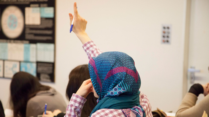 Ein Mädchen mit Kopftuch sitzt im Klassenzimmer und hebt die Hand.